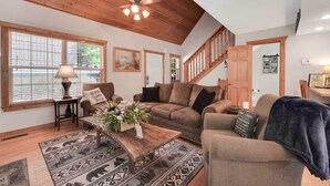 A spacious living room with a high ceiling, wooden beams, a large window with blinds, a sofa set, a coffee table with a floral arrangement, and a staircase leading to another level.
