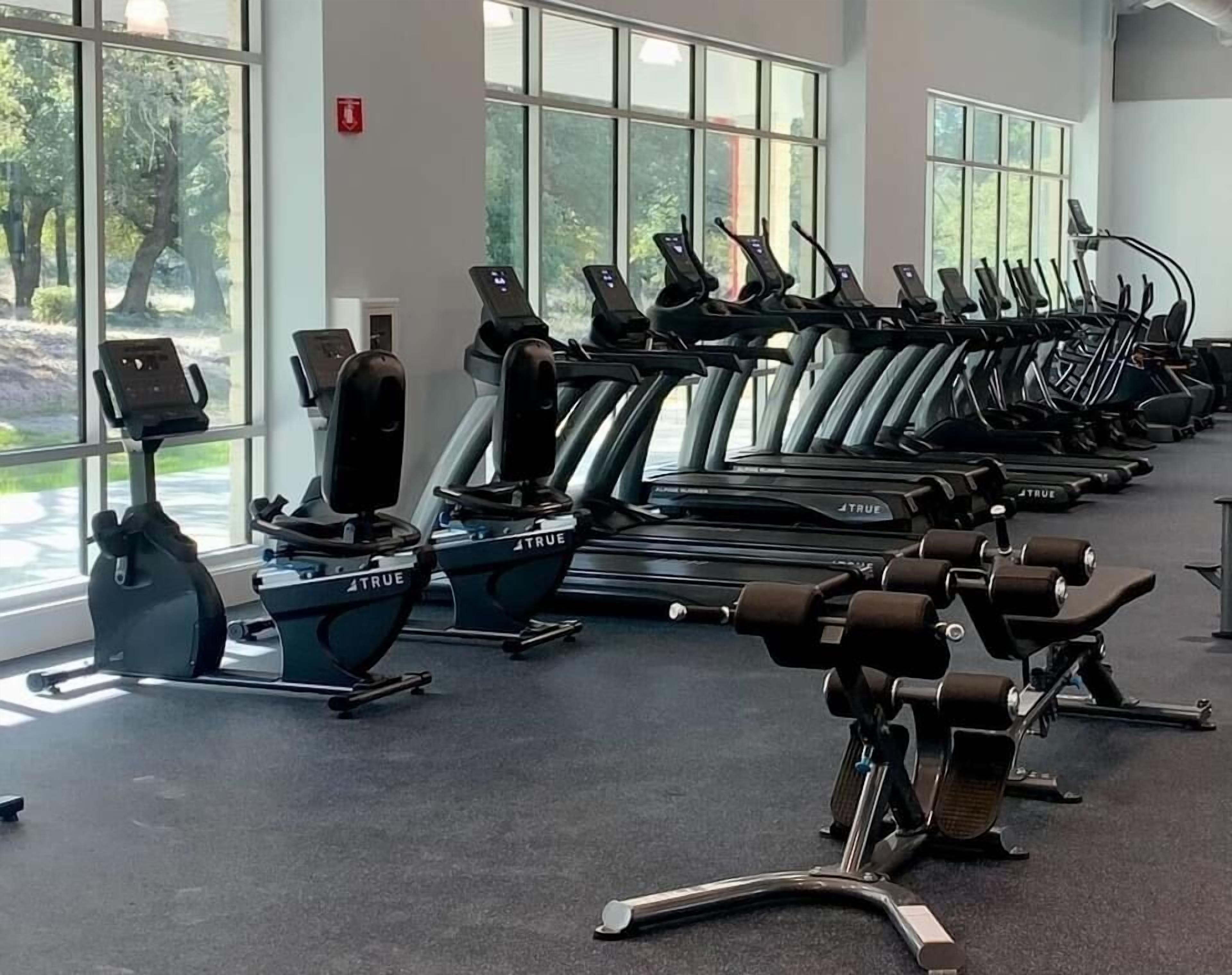 Ein Fitnessraum mit mehreren Laufbändern und Ergometer, großen Fenstern und einem Rauchmelder an der Wand.