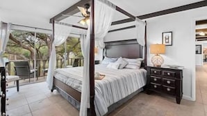 Uma cama com dossel e cortina, vista para árvores, mesa de cabeceira com abajur e uma cadeira em quarto com piso de azulejos.