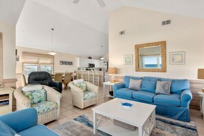 Ein geräumiges Wohnzimmer mit einem blauen Sofa, zwei Korbsesseln, einem weißen Couchtisch und einer Küche im Hintergrund.