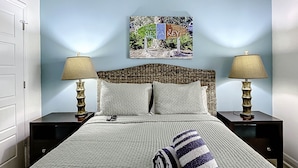A neatly made bed with a striped pillow, two bedside lamps, a framed welcome sign, and a wicker headboard. - The Beach Club at Siesta Key #106A Resort by RVA (Sarasota)