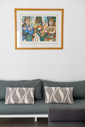 Een lijst met een abstract schilderij aan een muur, boven een grijze bank met gekleurde kussens.