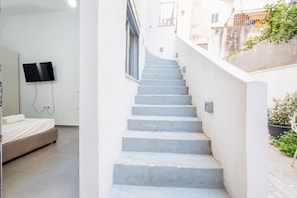 Un escalier mène à une chambre à coucher qui contient un lit, un téléviseur fixé au mur et une plante en pot dans l’espace extérieur.