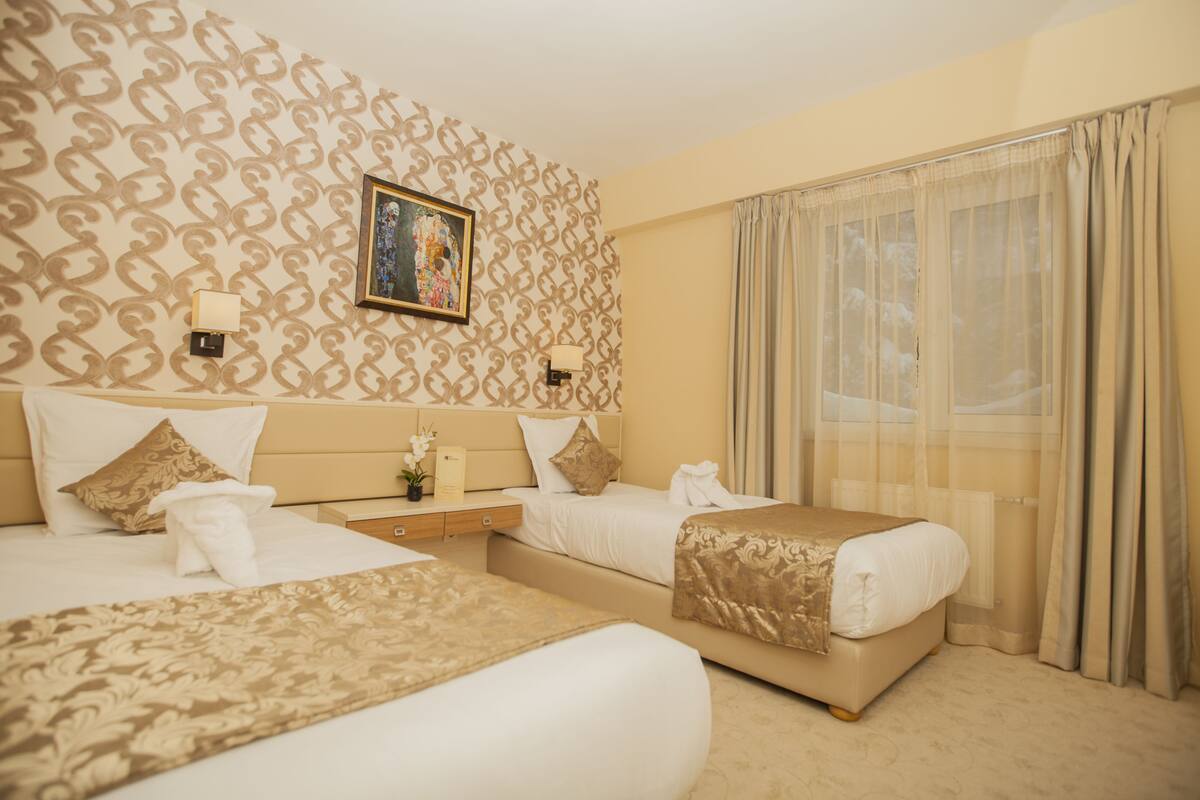 Een hotelkamer met twee bedden, een hoofdbord met een decoratief patroon, een lijst met kunstwerk erboven, een nachtkastje met een lamp, en een groot raam met gordijnen.