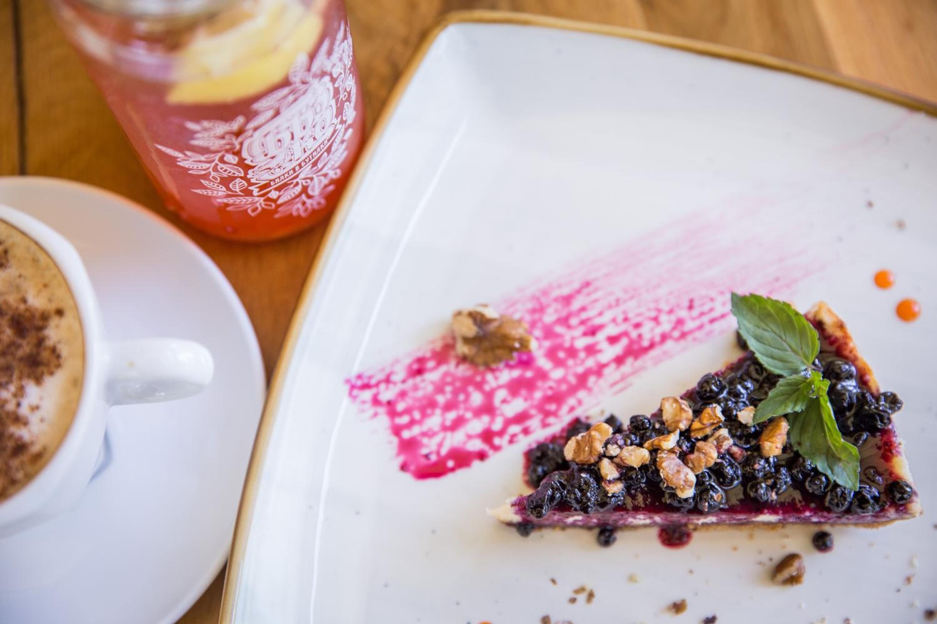 Une assiette à dessert avec une part de tarte aux myrtilles, garnie de noix et d’une feuille de menthe, accompagnée d’une tasse de café et d’un verre de boisson rouge agrémenté d’une tranche de citron.