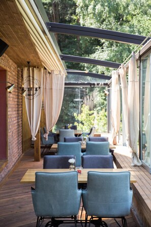 Un área de comedor al aire libre con mesas y sillas de madera, una pared de ladrillo y vistas a la vegetación.