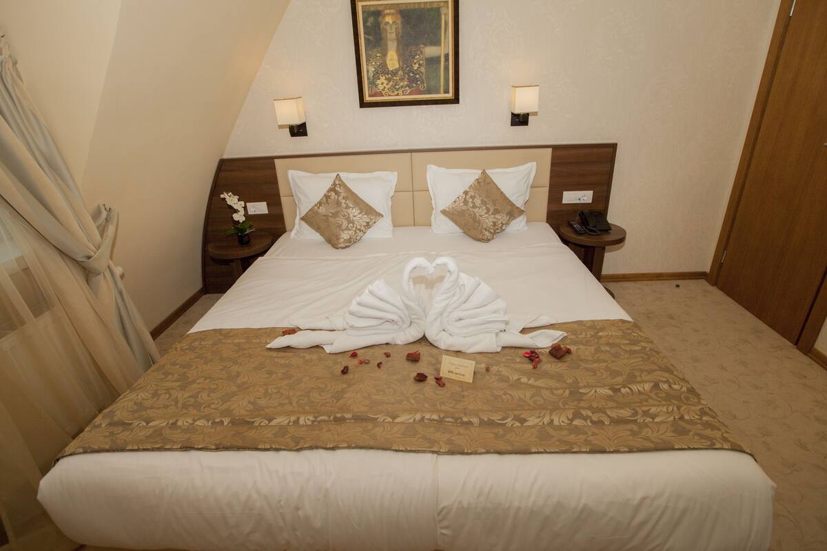 Een netjes opgemaakt bed met een decoratieve handdoekopstelling, een klein nachtkastje met een lamp, en een ingelijst beeld aan de muur.