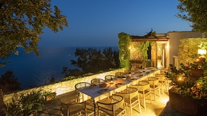 An outdoor dining area with a view of the sea, stone walls, and potted plants.