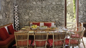 A dining area with a stone wall, a wooden table set for a meal, wicker chairs, and a red cushioned bench.