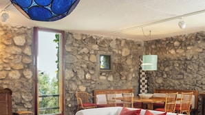 A living room with a stone wall, a glass coffee table, a white sofa with red cushions, and a dining area with wooden chairs.