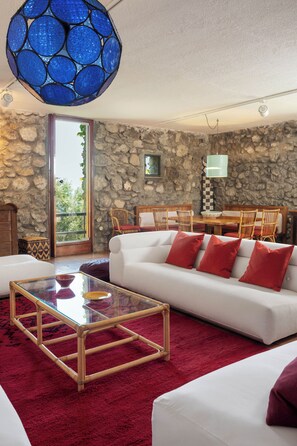 A living room with a stone wall, a white sofa, red cushions, a glass coffee table, and a blue hanging light fixture.