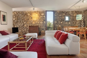 A living room with a stone wall, a white sofa with red cushions, a glass coffee table, and a dining area with a wooden table and chairs.