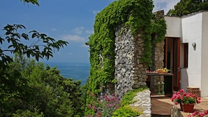 A stone building with a stone staircase leading to a door, surrounded by lush greenery and a view of the sea.