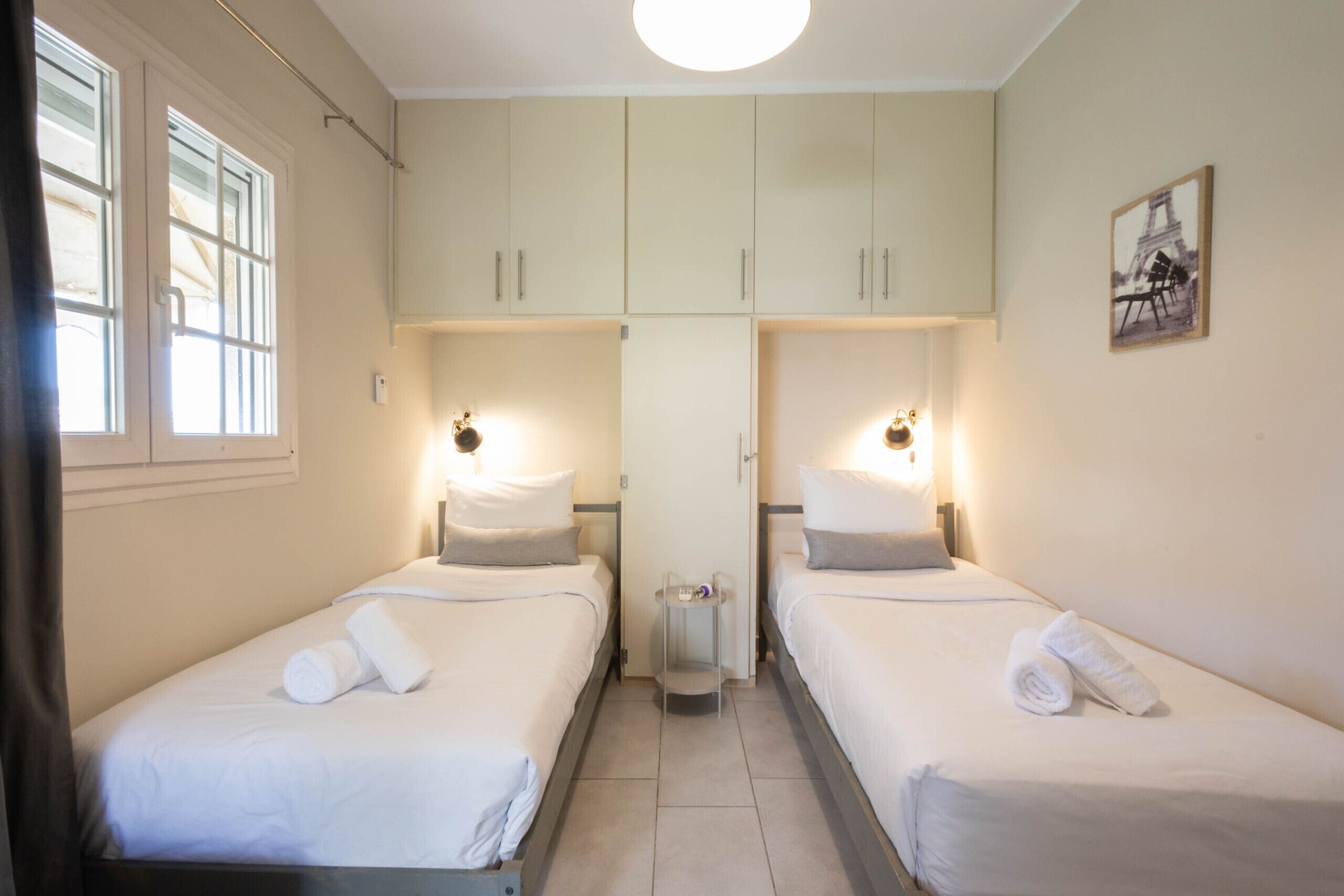 Ein Hotelzimmer mit zwei Einzelbetten, einem kleinen Tisch, einer an der Wand befestigten Ablage und einem Bild des Eiffelturms.