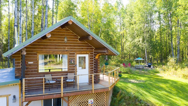 A log cabin with a balcony, a small deck, and a window overlooking a forested area. - Cozy Log Home -King bed, full kitchen, bathroom with clawfoot tub, outdoor area  (Talkeetna)