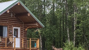 A log cabin with a balcony, a wooden deck, and a moose in the background. - Cozy Log Home -King bed, full kitchen, bathroom with clawfoot tub, outdoor area  (Talkeetna)