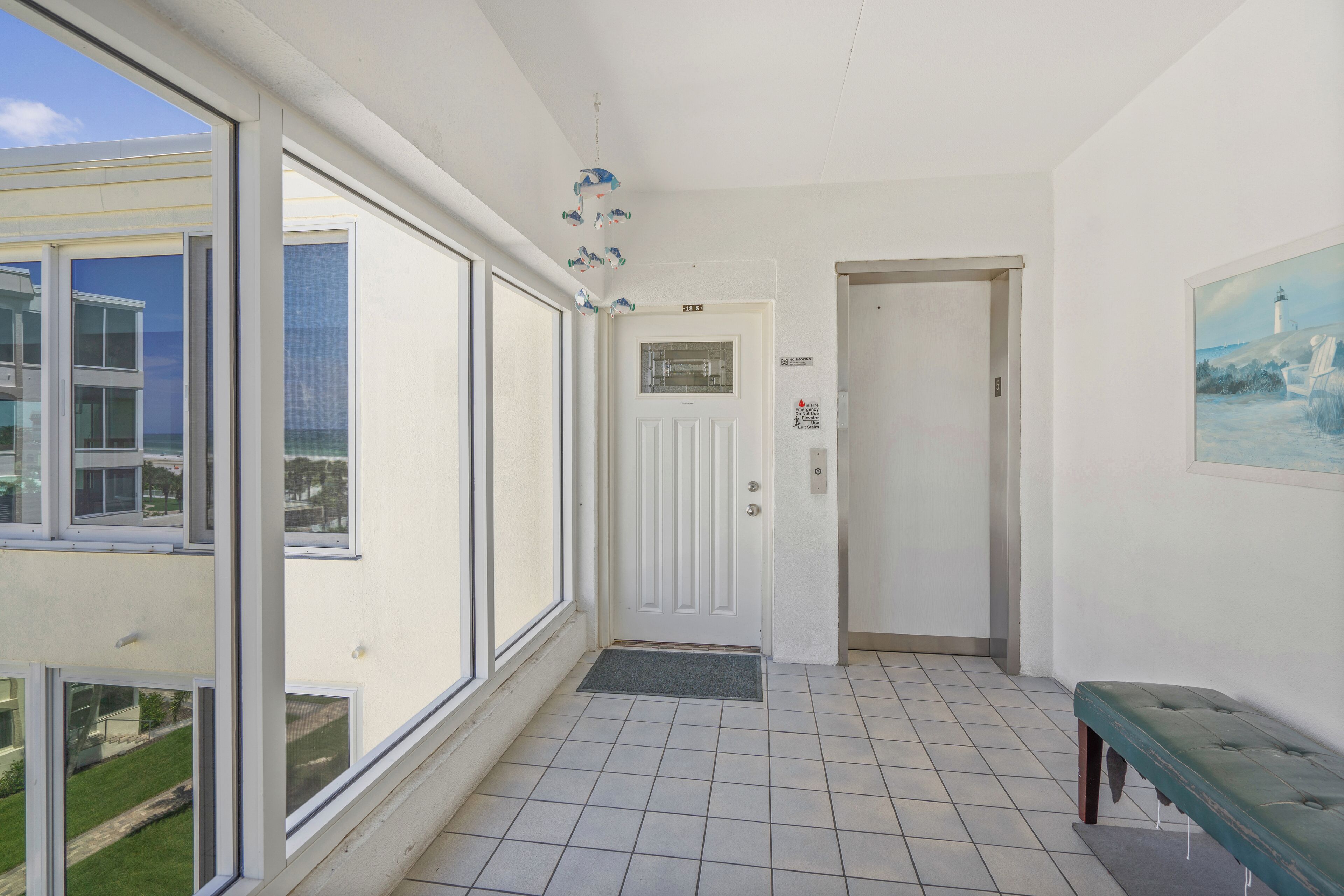 Een hotelkamer met een groot raam, een deur met een bordje, een bankje en een schilderij aan de muur.