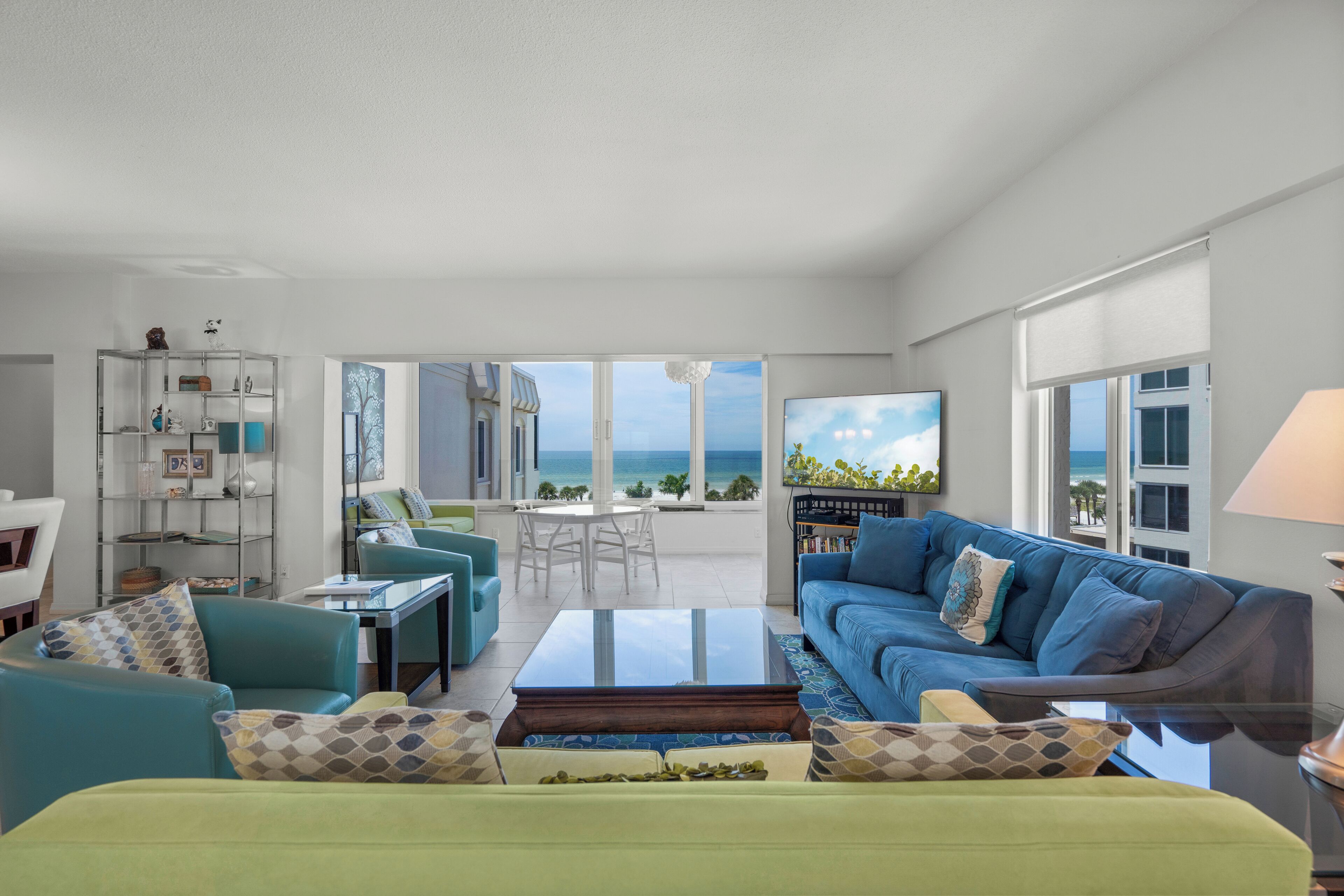 A spacious living room with a blue sofa, green armchairs, and a coffee table. A bookshelf and a dining area with a table and chairs are visible.