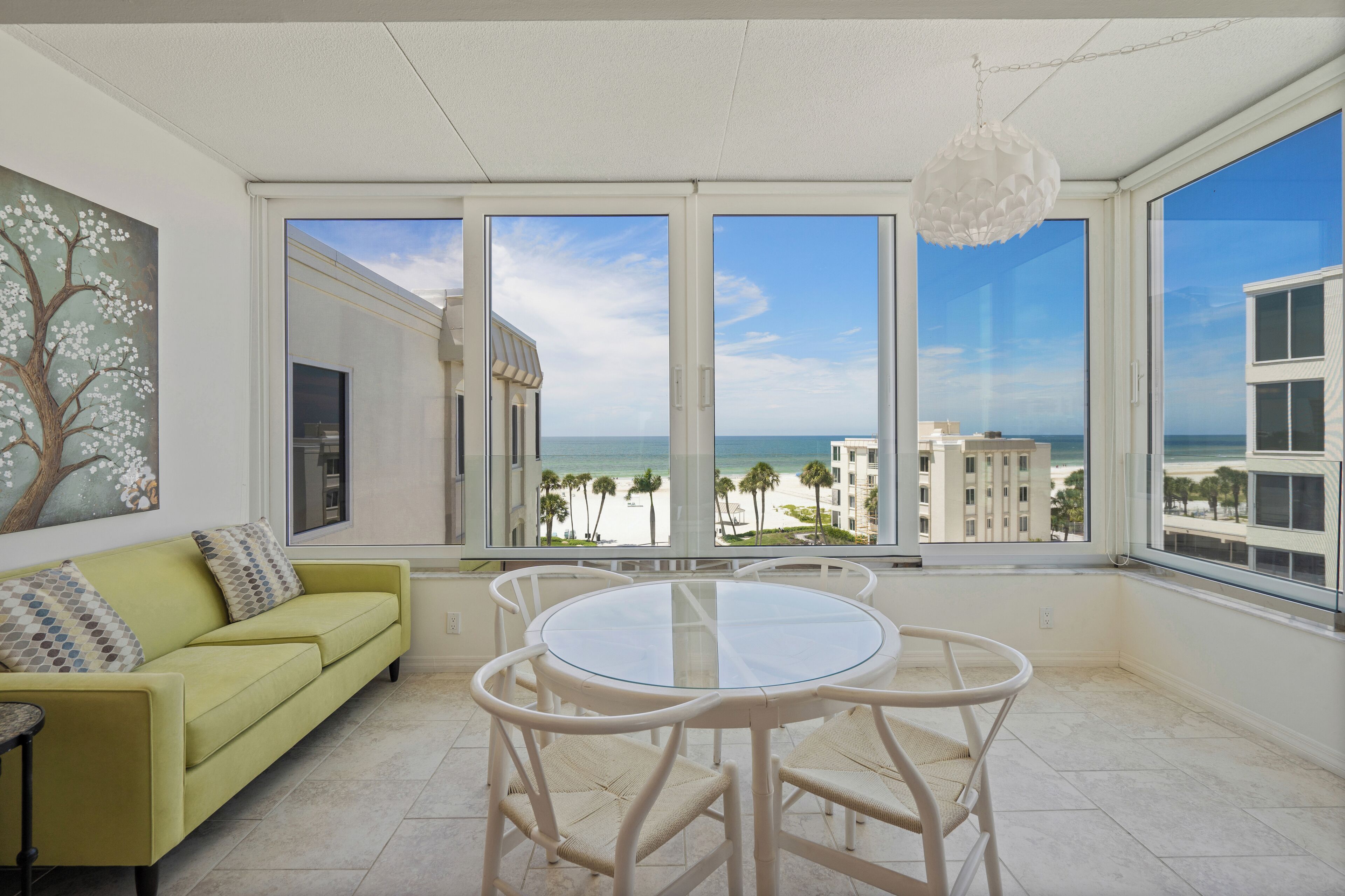 A room with a yellow sofa, a round glass table, and chairs, large windows with a view of the beach and ocean.