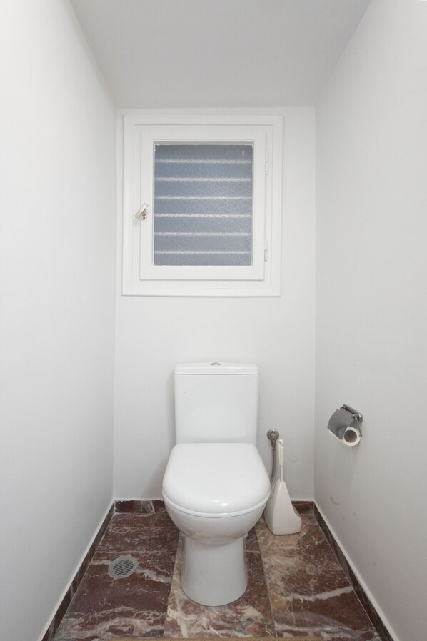 A small, white bathroom with a toilet, a bidet, and a window with blinds.