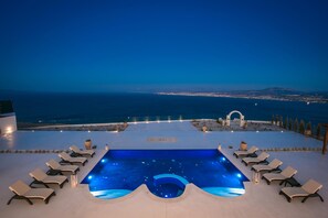 Une piscine à débordement située sur le toit, avec des chaises longues, une vue sur l’océan et un panorama de la ville en arrière-plan.