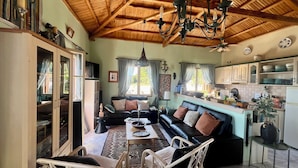 Ein gemütliches Wohnzimmer mit Holzbalkendecke, einer schwarzen Couch, einem Esstisch, Korbstühlen und einer Küchenzeile mit Schränken und einem Kühlschrank.