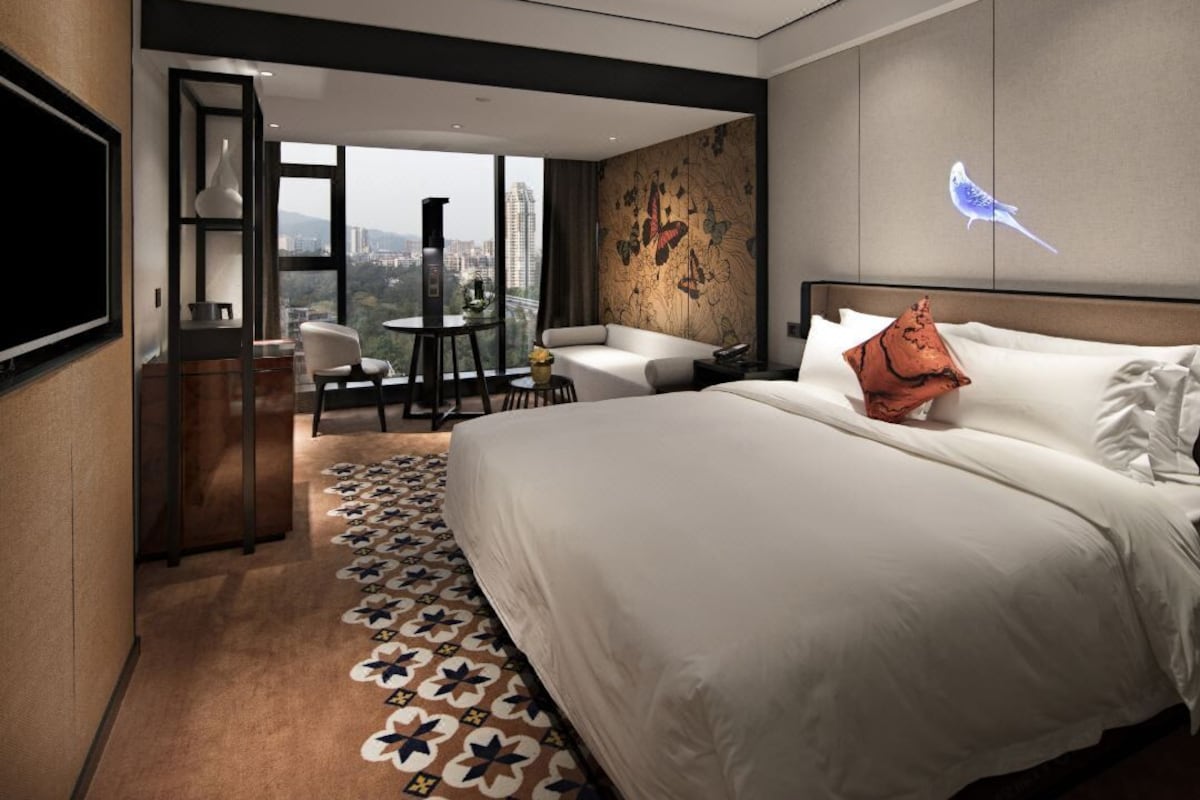 A modern hotel room with a large bed, a sofa, a dining table, and a city view.