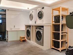 A laundry room with three stacked washing machines, a wooden shelf unit, and a small wooden stool. - Paco Hotel Zoo Metro Guangzhou (Guangzhou)