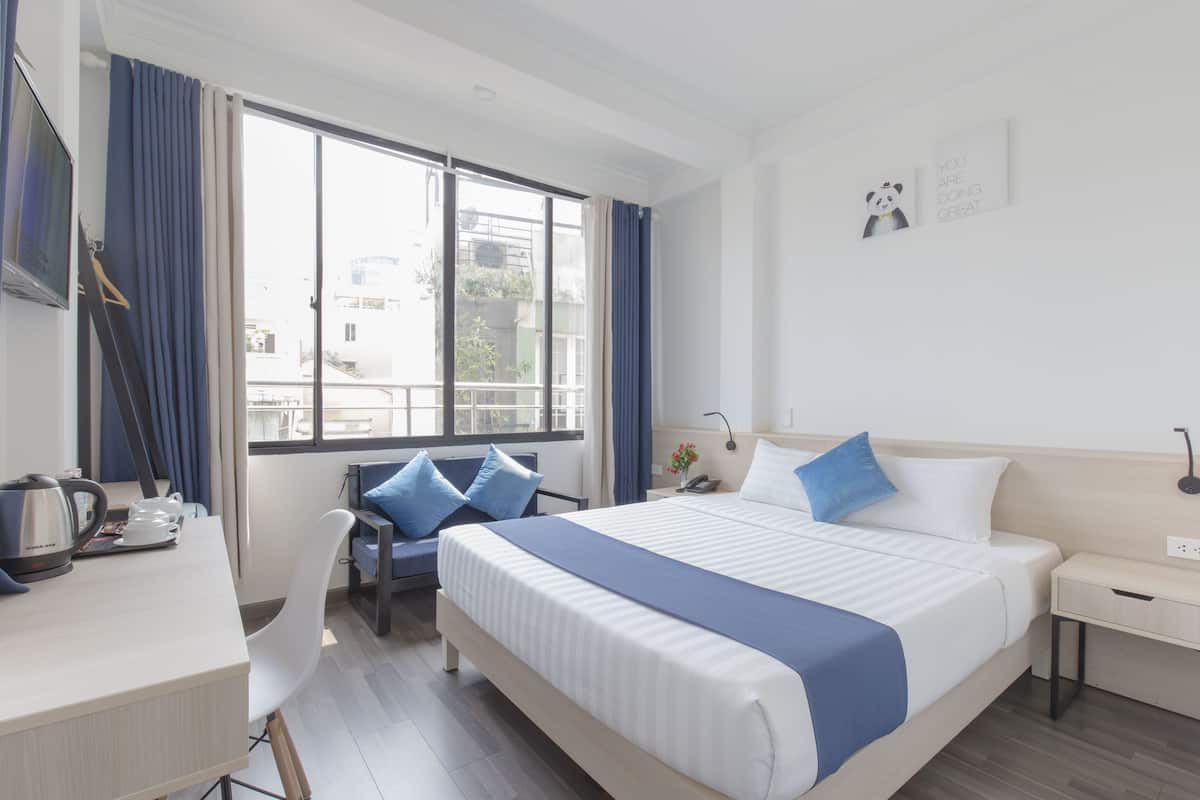 A modern hotel room with a large bed, a desk with a kettle, a chair, and a view of the city through the window.