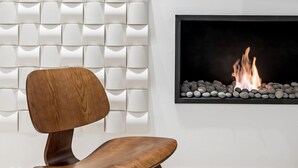 Ein modernes Interieur mit einer weißen, strukturierten Wand, einem flachbildfernseher über einem Kamin und einem Holzstuhl davor.