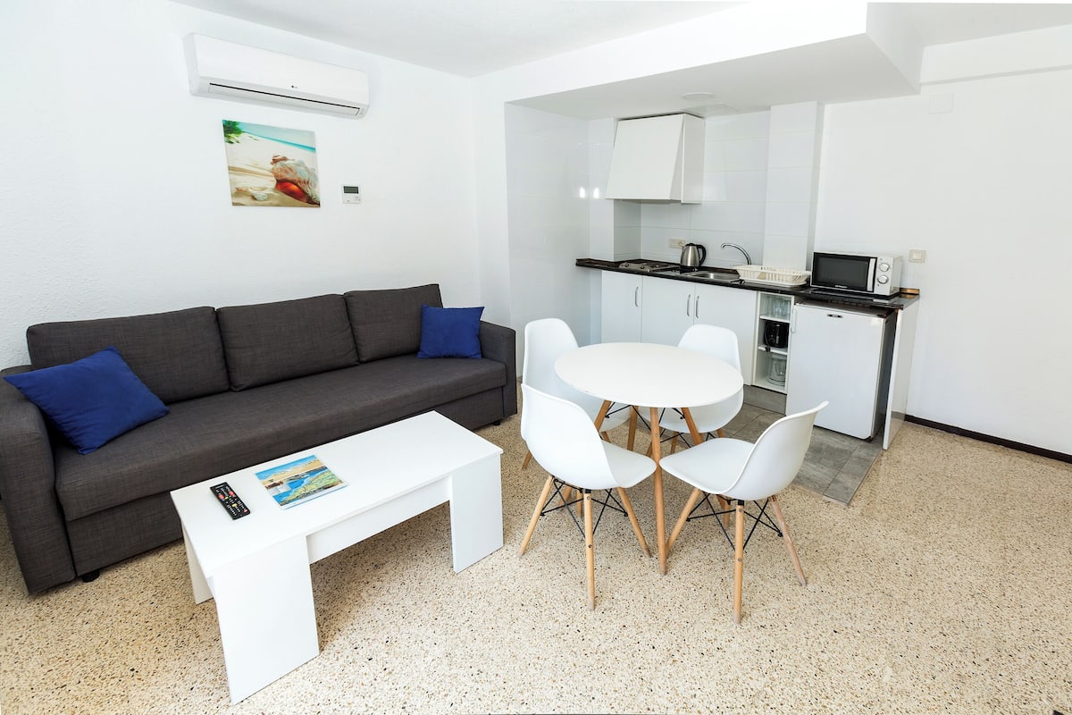 A modern living room with a grey sofa, white coffee table, and a small dining table with white chairs. A kitchen is visible in the background with white cabinets and a microwave.