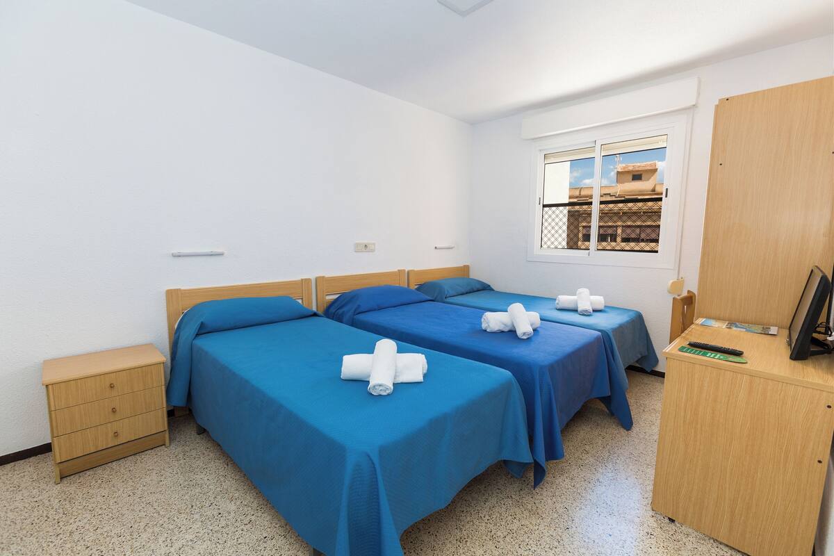 A room with two beds, a wooden desk, a small bedside table, and a window with a view of buildings.