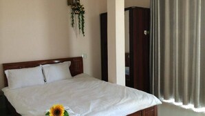 A neatly made bed with a white bedspread and a sunflower decoration, a wooden wardrobe, and a curtain on a window. - Nguyen Minh Hostel (Cam Ly - Da Lat)