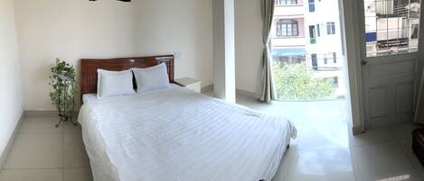 A hotel room with a large bed, a bedside table, a potted plant, and a view of the outside through a door.