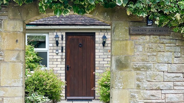 Une entrée en pierre avec une porte en bois, une enseigne indiquant « APPARTEMENT AVEC JARDIN », et une petite fenêtre avec un luminaire.