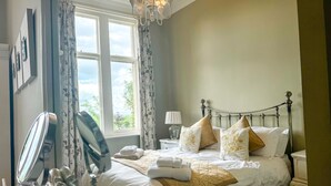 Un lit bien fait, avec une literie blanche et beige, une lampe de chevet et une vue sur l’extérieur par une fenêtre ornée de rideaux à motifs floraux.