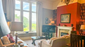 Un salon avec un canapé, une table basse, un téléviseur et une cheminée. On y trouve également des étagères et un lustre.