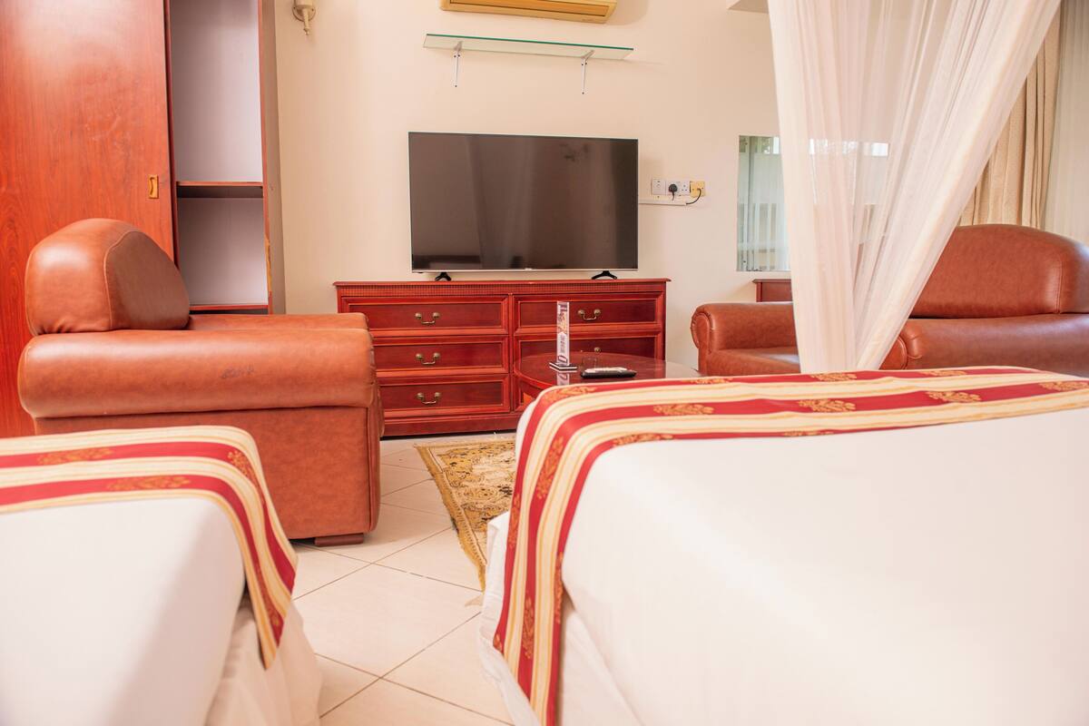 Habitación de hotel con cama, dos sillones, cómoda de madera, televisor y ventana con cortinas.