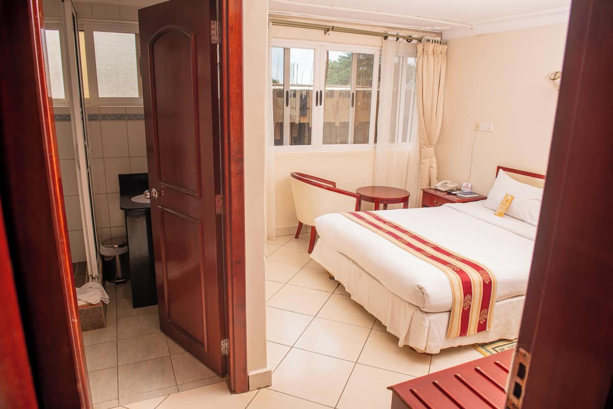Habitación de hotel con cama, una mesita, una silla, ventana con cortinas y baño con inodoro y lavamanos.