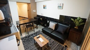 Uma sala de estar moderna com sofá de couro preto, mesa de centro de madeira, tapete estampado, mesa de jantar com cadeiras e área de cozinha com micro-ondas.