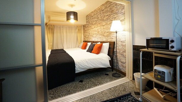 Quarto de hotel com cama, luminária, micro-ondas e uma pequena prateleira com torradeira.