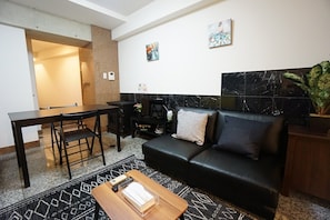 A modern living room with a black leather sofa, a wooden coffee table, a dining table with chairs, and a small TV.