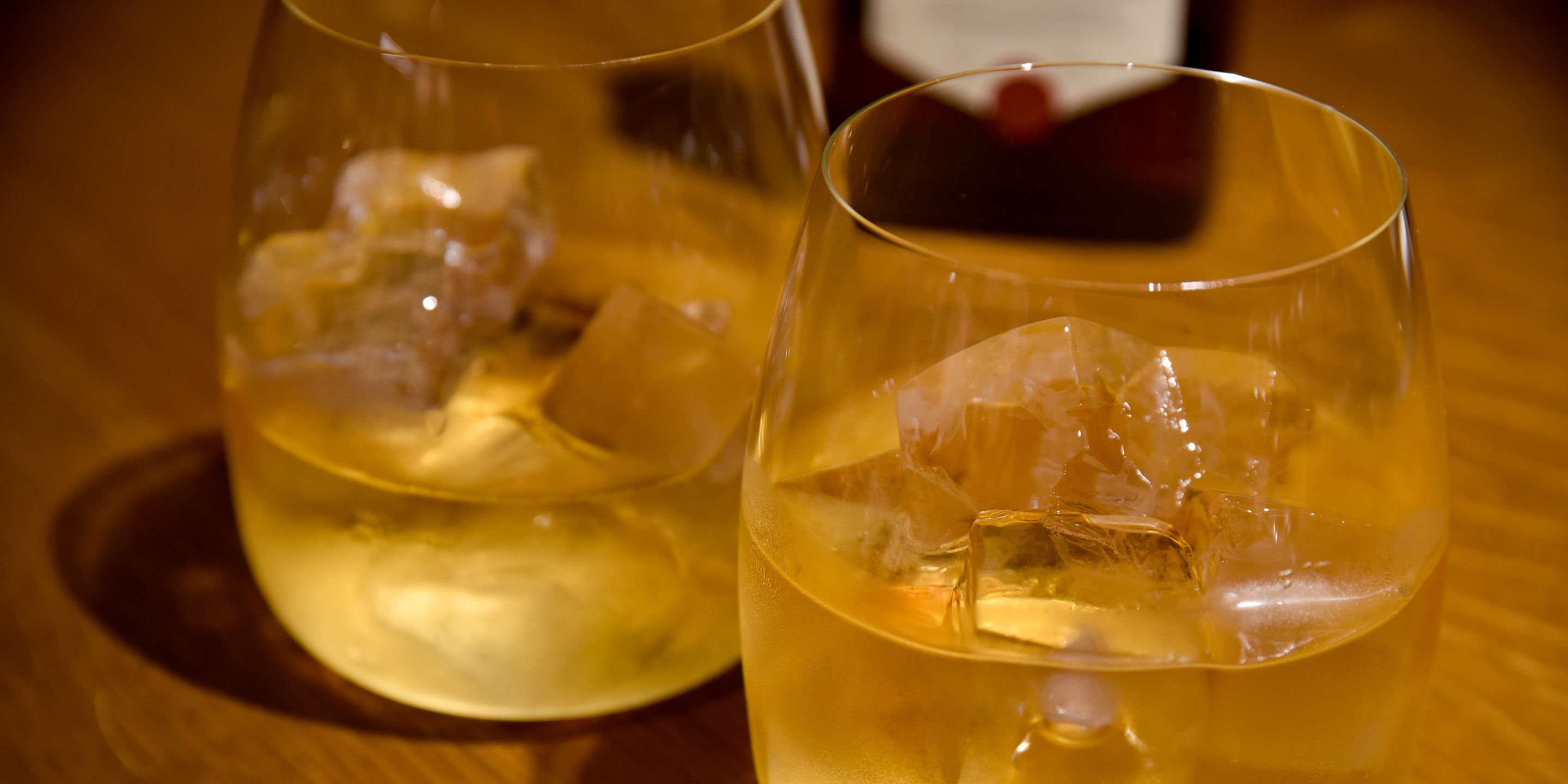 Dos vasos de bebida color ámbar con cubitos de hielo sobre una superficie de madera.