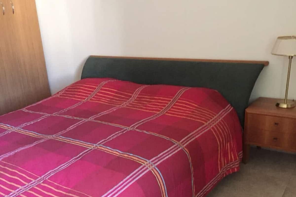 A bed with a red and green bedspread, a wooden headboard, a nightstand with a lamp, and a tiled floor.