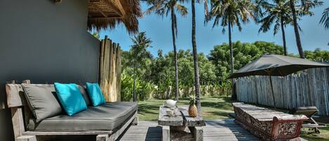 A wooden deck with a sofa and a table, surrounded by palm trees and a thatched-roof structure.