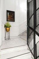 Uma escadaria com degraus de mármore branco, corrimão de metal preto e uma planta em vaso sobre um suporte dourado.