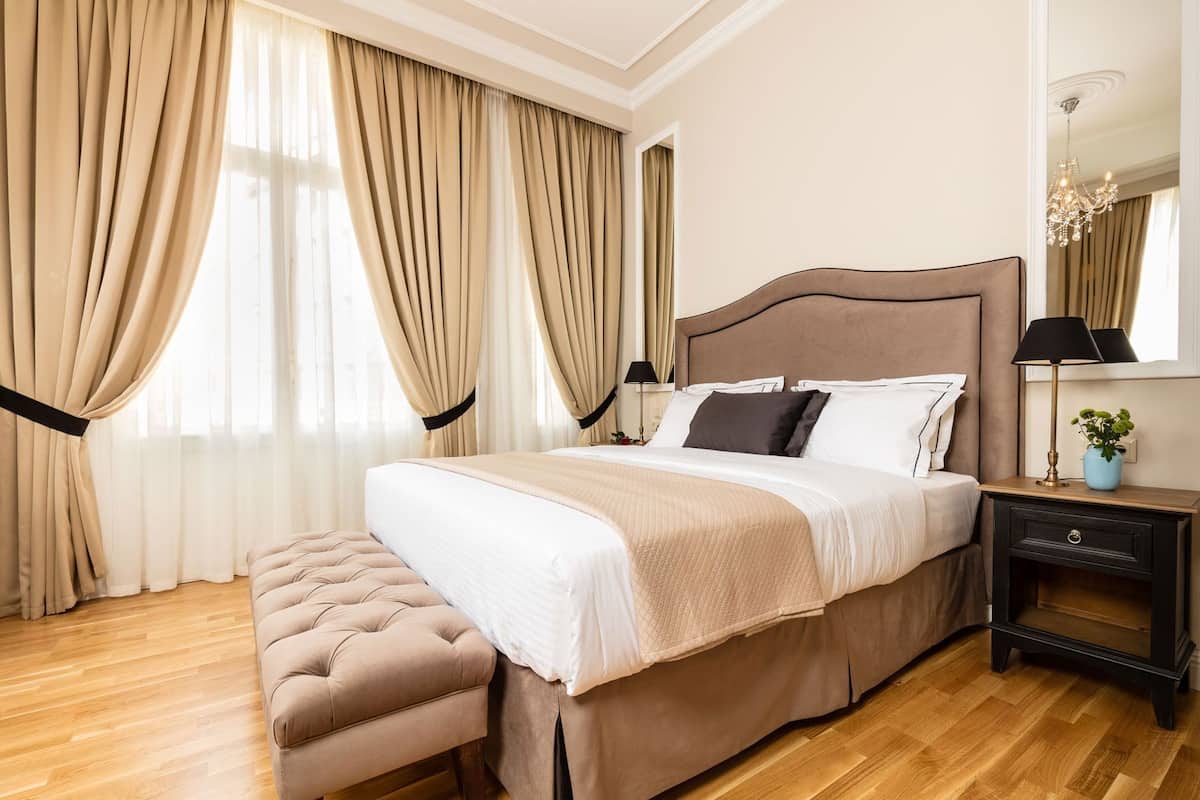 A neatly arranged bedroom with a large bed, a bedside table with a lamp, a bench, and a window with curtains.