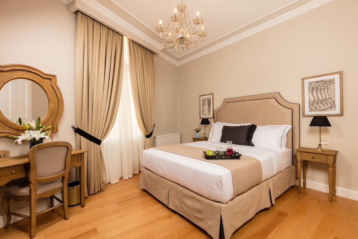 A hotel room with a bed, a desk, a chair, a chandelier, and a window with curtains.