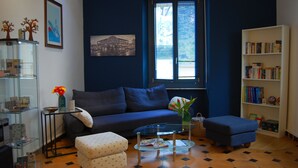 A living room with a blue sofa, a glass coffee table, a bookshelf, and a window with blinds. - Colazione da Augusta (Turin)