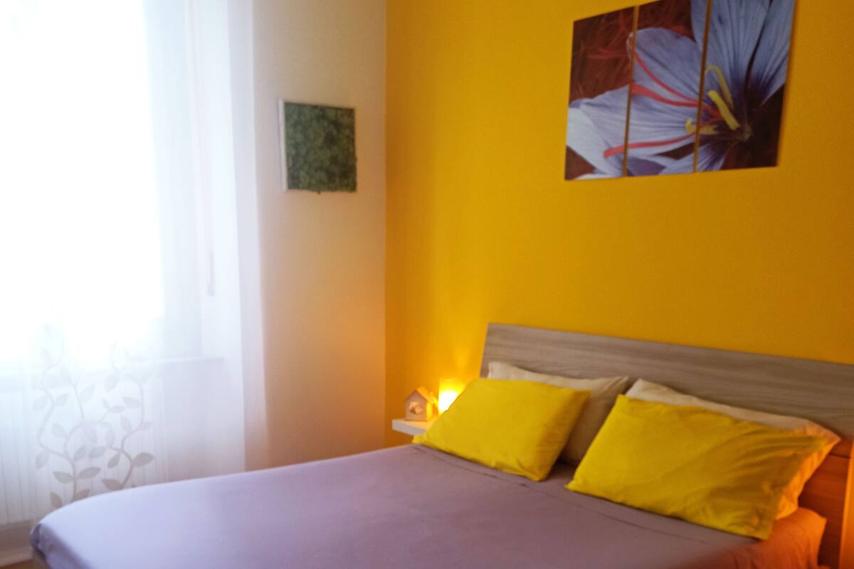 A bed with a purple bedspread and two yellow pillows, a nightstand with a lamp, and a wall with a framed picture.